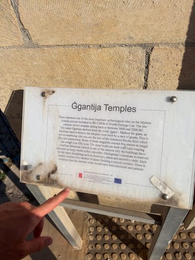 A signpost with information about the Ggantija Temples is displayed outdoors. The sign is made of a white material and is covered in dirt and smudges. A person's hand is visible pointing at the sign. The background consists of a stone wall and a metal stand supporting the sign. The stone wall is made of large, rough-hewn blocks. The ground is covered in a pattern of circular indentations. The scene appears to be in Xagħra, Malta, as indicated by the sign's text which references the Ggantija Temples.