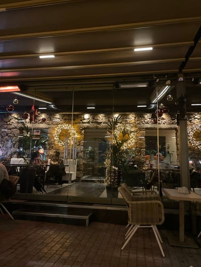 This image depicts an outdoor or semi-outdoor seating area of a restaurant or cafe in Chaidari, Greece, at night. The scene is brightly illuminated by artificial lighting, creating a warm and inviting atmosphere.
The foreground and right side of the image show an outdoor paved area, featuring light-colored brickwork. Several chairs, some stacked on tables, are visible, suggesting it might be past operating hours or preparing to close. A glass partition separates this outdoor space from the interior of the establishment.
On the left side and overhead, there is a retractable awning or roof structure with embedded strip lighting that casts a bright glow. Infrared heaters are mounted to this structure, suggesting the outdoor area is designed for comfort during cooler evenings.
Beyond the glass partition, the interior is distinctly visible, featuring a prominent, textured wall made of stones or rocks. This wall is extensively decorated with numerous warm-toned fairy lights, some arranged into decorative wreaths. Green foliage and potted plants, also adorned with lights, are interspersed along the wall. Several tables and chairs are arranged inside, and a few patrons are faintly visible, occupying some of the seating.
The overall impression is one of a festive and cozy venue, likely catering to evening diners or social gatherings. No discernable text is legible within the image.