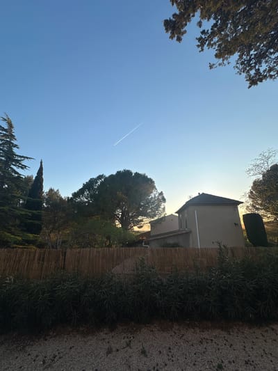 The image shows a view in Lourmarin, France, with a building visible against a clear blue sky. A contrail from an airplane is visible in the sky. Trees frame the view on both sides. A wooden fence and dense foliage are present on the right side of the frame, along with a gravel path.
