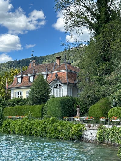 A grand, yellow-sided house with a red tiled roof stands prominently by a body of water. The house features multiple dormer windows, a large arched window, and several chimneys. It is surrounded by lush green trees and manicured hedges, with a stone wall and a metal fence lining the waterfront. Red flowers in white planters add pops of color along the fence. The water is a clear, turquoise blue, reflecting the bright, cloudy sky above. The scene is set against a backdrop of verdant, tree-covered hills. The house appears to be located in the vicinity of Hünibach, Switzerland, given the characteristic Swiss landscape and architecture.