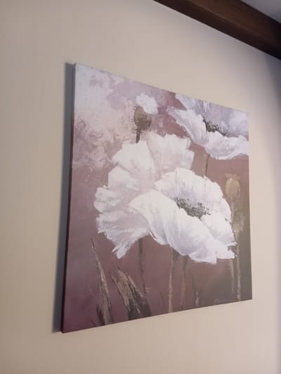 An abstract painting of white poppies hangs on a plain, beige wall. The painting is rectangular and oriented horizontally. The poppies are rendered with thick, brushstroke textures, predominantly in white and pale pink, against a muted, dusty rose background. Several tall, thin green stalks are visible in the foreground. A dark brown wooden beam runs horizontally across the top of the frame, casting a shadow on the wall above the painting. The painting is signed in the bottom right corner, though the signature is illegible. The scene is indoors, likely in a domestic or gallery setting. The location in Baden, Austria, is not specified by any visual cues in the image.