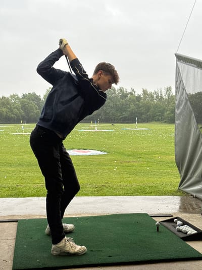 A young man wearing a black sweatshirt, black pants, and white sneakers is swinging a golf club at a golf ball on a green mat. He is standing on a concrete pad, and there is a gray netting behind him. There is a green grassy area beyond the concrete pad, and there are white golf balls scattered across the grass. There are also several white tee markers in the distance. The image is taken at a golf driving range located in South Ribble, United Kingdom, at the coordinates 53.771, -2.57. The weather appears to be overcast, and there is a light mist or drizzle falling. The man is focused on the golf ball, and his face is obscured by the brim of his hat. He is wearing a white golf glove on his left hand, and his right hand is gripping the club. The golf club is swung with force, and the ball is in motion. The man is positioned to the left of the frame, and the golf driving range is to the right of the frame. The overall scene is one of concentration and focus. The man is practicing his swing, and the image captures the moment of impact between the club and the ball.