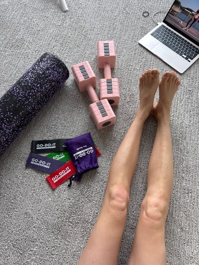 The scene is an indoor, overhead view in Springfield, United States of America, showing a person's bare lower legs and feet extended on a gray textured carpet. To the upper left, a black foam roller with purple speckled patterns rests horizontally. Centrally positioned are two pink adjustable dumbbells, each featuring gray weight adjustment mechanisms. Below these dumbbells, a collection of "GO-DO-IT Fitness" resistance bands in black, green, purple, and red are laid out, alongside a purple "GO-DO-IT Pro Bands" drawstring pouch. To the upper right, a silver laptop is open, displaying a fitness video on its screen that shows an individual in athletic wear outdoors. A black hair tie is visible near the top left corner of the laptop, and a white cable partially enters the frame from the top right.