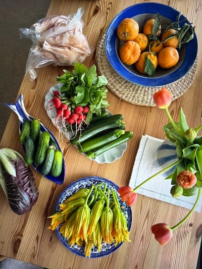 An overhead view captures a vibrant array of fresh produce and flowers arranged on a light wooden table.

In the upper left, a blue bowl holds several oranges, some still bearing green leaves, resting on a round woven straw placemat. Below this, a whole raw chicken, still in its clear plastic packaging, is visible. Adjacent to the chicken, a bunch of red radishes with their green tops is placed next to multiple green zucchinis in a white scalloped dish.

Further down on the table, a distinctive blue and white fish-shaped dish contains several small cucumbers. To its right, a head of purple radicchio or red cabbage lies on the wooden surface.

In the upper center of the frame, a blue and white striped vase holds a bouquet of green-stemmed tulips. Some tulips are open, displaying orange-red petals, while others remain in bud. The vase sits on a folded white cloth. To the right of the flowers, a blue and white patterned plate is filled with numerous pale yellow-orange squash blossoms.

The scene depicts a rich collection of fresh ingredients, suggesting preparation for a meal. The lighting indicates daytime. There are no discernible location cues or visible text within the image.