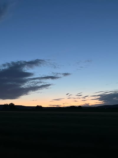 The image shows a view of the sky and a dark landscape, presumably taken near Gorna Kremena, Bulgaria. The sky transitions from a deep blue on the left to a lighter, orange-tinged hue towards the right horizon. Several dark cloud formations are visible in the lower center and towards the upper right of the sky. A dark landmass, possibly a field or forest, forms the right edge of the image, with some trees distinguishable against the sky. The image is taken at twilight.