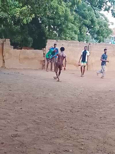Five young men are playing soccer on a dirt field in what appears to be a residential area in Oke Ogun, Nigeria. A crumbling, low wall surrounds the field, and lush green trees are in the background. One of the men is actively kicking a soccer ball; he is shirtless and wearing shorts. The others are wearing various attire, including jerseys and casual clothes. One man is walking away from the group. The environment is dusty and somewhat barren, with scattered debris on the ground.