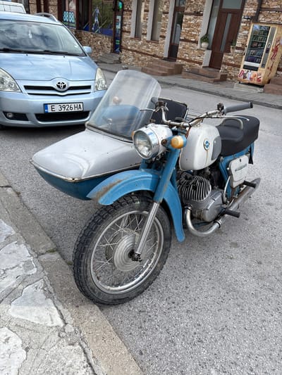 The image depicts a vintage blue and white BMW motorcycle with a sidecar parked next to a silver Toyota Corolla in the city of Bachevo, Bulgaria. The car has the Bulgarian license plate E 3926 HA. The motorcycle is parked on asphalt pavement near a sidewalk curb. In the background, there is a brick building with a vending machine and a shop with a glass storefront. The angle of the photo suggests it was taken from a standing position. There are no visible people in the frame.