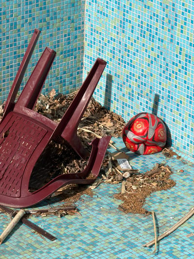 The image depicts an abandoned swimming pool in Nerin, Montenegro. The pool is tiled with blue and green mosaic squares and is littered with debris, including dry leaves, twigs, and a partially broken brown plastic chair. A red and grey football with the Manchester United logo rests near the pool's edge. There are no people visible in the scene.