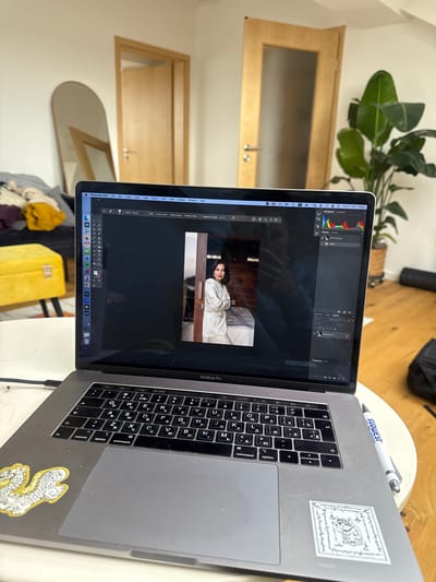 A laptop is open and running Adobe Photoshop 2026. The image on the screen shows a woman in an indoor setting. The laptop keyboard shows the letters in Russian. There is a Southwest pen, and a sticker on the laptop. The laptop sits on a desk, and there is a plant and a mirror in the background. The laptop is most likely a MacBook Pro. There is no information about the city of Ampfing, Germany.