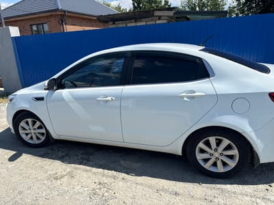 The image shows a white Kia sedan parked on a gravel road in front of a blue metal fence. A brick house with a dark roof is visible behind the fence. The car is viewed from the side, with the front passenger door and rear passenger door visible. The windows are tinted. The environment appears to be a rural or suburban area. The time of day is daytime, as indicated by the shadows cast by the car. There are no people visible in the image.