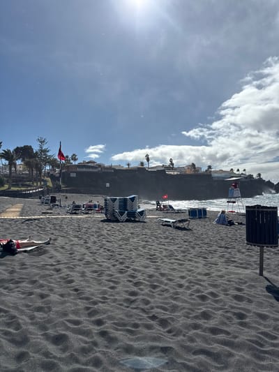 This image depicts a daytime beach scene at Puerto De Santiago, Spain, characterized by black volcanic sand. The sun is high, causing a bright glare in the upper left sky, which is partly cloudy with patches of blue.

In the foreground, the rippled black sand dominates the lower half of the frame. On the far left, an individual lies supine on a red towel directly on the sand, dressed in red swimwear, with a black bag near their head. To the right, a dark grey cylindrical public waste bin on a pole stands.

In the mid-ground, a large stack of approximately 20-25 light blue and white plastic sun loungers is centrally positioned. Further right, closer to the water, a white lifeguard chair with red accents and a red flag attached stands. An individual is seated on a sun lounger near the lifeguard chair, partially covered by a blue and white striped towel, facing the ocean. A smaller red flag on a pole is visible near the water's edge, between the stacked loungers and the lifeguard chair. A wooden boardwalk or path extends from the left side of the frame towards the mid-ground, partially covered by sand. In the background, beyond the beach, the ocean features visible waves breaking against the shore or rocks.

The background consists of a dark, rocky coastline or cliff face, topped with white residential buildings, houses, and apartment complexes. Numerous palm trees and other green foliage are interspersed among the buildings and along the coastline. Another tall red flag on a pole is visible on the far left, above the boardwalk.