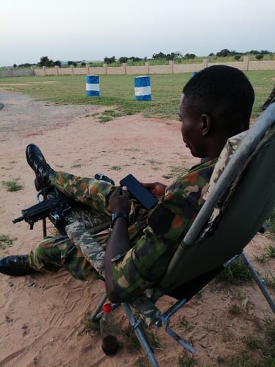 In Kayauki, Nigeria, an outdoor scene depicts a male individual, likely a soldier, seated in a collapsible olive green chair on reddish-brown earth with sparse grass. He is wearing a camouflage uniform (consistent with Nigerian Army pattern), black tactical boots, and a black digital watch on his left wrist. His legs are extended, resting over an assault rifle, which appears to be a bullpup design (e.g., IWI Tavor variant), with its barrel pointing left. He is actively engaged with a black smartphone, holding it with both hands and looking down at its screen. A clear plastic bottle with a red cap, containing a dark liquid, rests on the ground near the base of his chair. In the background, a large grassy field extends, featuring two blue and white striped barrels positioned as potential targets or markers. Beyond the field, a low concrete block wall is visible, with scattered trees and bushes behind it. The sky above is light, suggesting either early morning or late afternoon conditions.