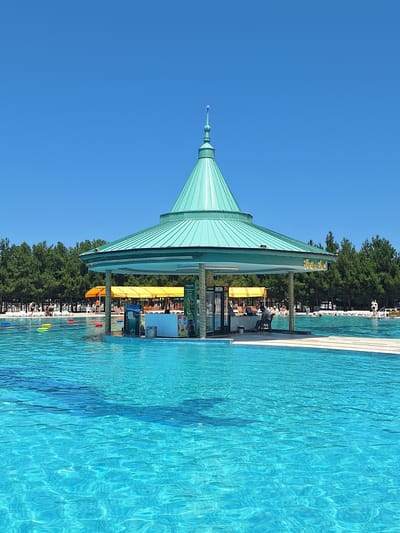 The image shows a bright, sunny day at a swimming pool in Khudat, Azerbaijan. In the center of the frame is a circular bar with a green, conical roof, situated directly in the pool. The bar area is open with people working behind it and patrons sitting at it. The pool water is a vibrant turquoise color with visible ripples and reflections. Around the pool, there are lounge chairs and yellow umbrellas, with a line of green trees forming the background. Several people are visible in and around the pool, some swimming and others relaxing.