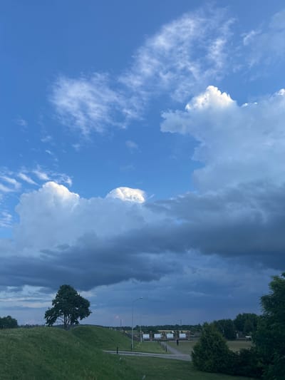 This image depicts a landscape under a dramatic sky, likely in or near the village of Zhuhavichy, within the Zhukhavichy rural council of Belarus. The foreground features a grassy embankment with a lone, mature tree on its crest, casting a subtle shadow. To the right, a paved pathway runs alongside the embankment, bordered by low bollard lights and a few small, manicured evergreen bushes. Further into the midground, a series of white gates are visible, along with a solitary figure walking along the pathway. The background is a tapestry of trees, forming a dense line on the horizon. The sky dominates the upper portion of the frame, characterized by a striking contrast between bright, fluffy white cumulus clouds illuminated by the sun on the left and darker, heavy storm clouds gathering on the right, creating a sense of impending weather change. The lighting suggests it is either late afternoon or early evening.