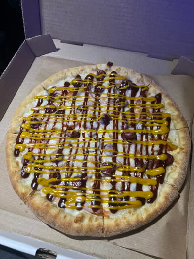 A whole pizza is presented in a cardboard pizza box. The pizza is topped with cheese, sliced sausages, red onions, barbecue sauce, and mustard. The box is positioned on a brown surface, likely the interior of the box. The background is dark, suggesting a low-light environment, possibly at night or indoors. There is no indication of the city of Bakaly, Russia in the image.