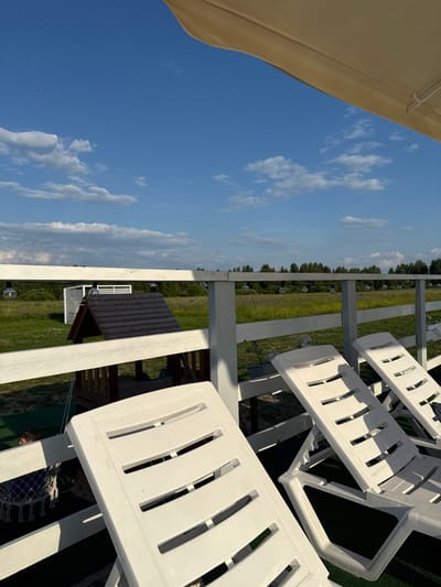 The image shows a clear blue sky with scattered white clouds, with a section of a beige umbrella in the upper right corner. Below the sky, there is a white wooden fence, and beyond it, a green field with trees in the distance. In the foreground, there are several white plastic lounge chairs. To the left of the lounge chairs, a small wooden structure with a dark roof is visible, resembling a playhouse or gazebo. A young boy is partially visible behind the fence, sitting in a white netted swing. The scene appears to be an outdoor recreational area, possibly a resort or a park, located in or near Koloksha, Russia, given the context of the prompt. The lighting suggests it is daytime, likely late afternoon or early evening due to the long shadows.