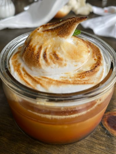 A jar of dessert sits on a wooden surface, featuring a toasted meringue topping with a swirl pattern and a single green leaf as garnish. The jar reveals a caramel-colored filling underneath. White napkins and small bread rolls are visible in the blurred background, adding to the dessert presentation. The jar is slightly out of focus on the side that is farthest away.