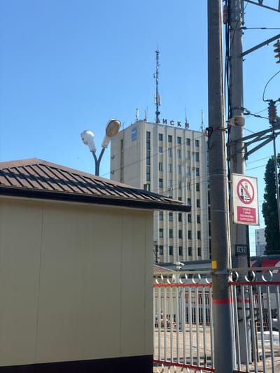 A sunny, outdoor, daytime scene captures a view of a tall, light-colored building with red lettering spelling "ЛИCKИ" on top. The building has numerous windows and is topped with various antennas. In the foreground, a brown metal-roofed structure partially obscures the view. To the right, a concrete utility pole is adorned with a white sign featuring a red circle with a diagonal line through it, depicting a person standing near a train track. Below this symbol, red Cyrillic text reads "СТОЯТЬ У КРАЯ ПЛАТФОРМЫ ЗАПРЕЩЕНО!" which translates to "Standing at the edge of the platform is prohibited!". A metal fence with red and silver accents separates the foreground from a train track where a train is visible. The sky is a clear, bright blue. The image was likely taken at a train station in Zaluzhnoye, Russia, given the presence of the station-related signage.