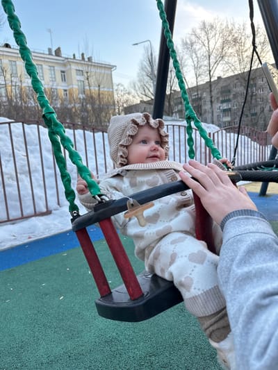 An infant, dressed in a light beige knitted bonnet and matching heart-patterned outfit with a pacifier clip, is seated in a red and black baby swing. An adult's fair-skinned hand rests gently on the swing's front bar, while another hand, partially visible, holds a light-colored smartphone to the right, possibly documenting the scene. The swing chains are covered in green plastic. The ground beneath the swing is a multi-colored rubberized playground surface, primarily green and blue. Beyond the playground, snow is visible on the ground, bordered by a low white fence. In the background, several multi-story residential buildings stand under a clear blue sky, with bare deciduous trees scattered between them. This scene is located in a city environment in Sadovaya, Russia, during what appears to be late winter or early spring.