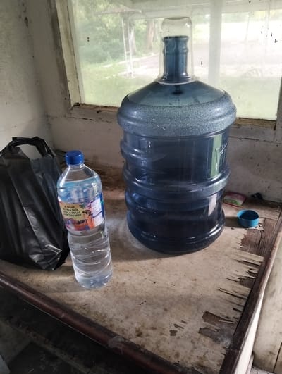 In Palohgadeng, Indonesia, a close-up shot depicts two large, blue, plastic water jugs placed on a weathered wooden table in front of a grimy, frosted window. The larger jug, situated to the right, is topped with a clear plastic cup acting as a temporary seal. To its left, a smaller, clear plastic bottle of water stands upright. Behind the smaller water bottle, a crumpled black plastic bag rests against a white wall. The surface of the table is marred by water stains and peeling veneer. Through the window, blurry green foliage and the indistinct structure of a building are visible.