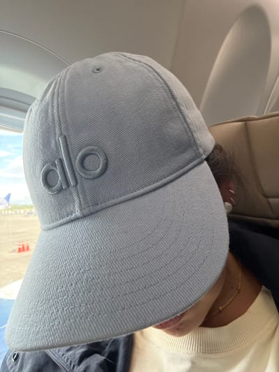 The image depicts a person wearing a light gray Alo cap, seemingly on an airplane. The plane window and parts of the airport are visible. The cap's brim is textured. The seat headrest and the person's light colored sweater and a gold necklace are visible, with glimpses of their face near the brim of the hat.