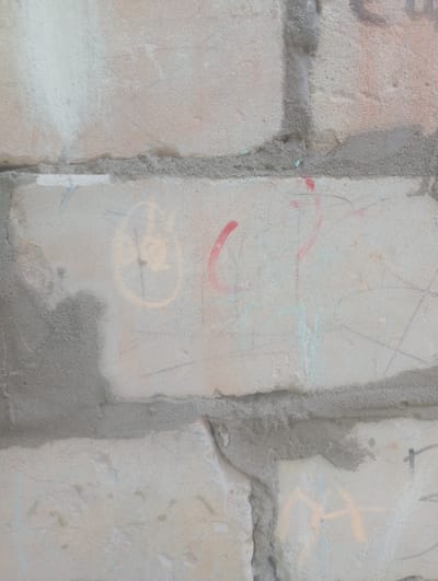 This media file is a close-up, eye-level shot of a brick wall with some graffiti. The wall is made of light-colored bricks and has visible mortar lines. The graffiti consists of various scribbles and shapes in red, yellow, and light blue. Some of the markings are faint and partially obscured by the texture of the wall. There are no people or objects clearly depicted. The scene appears to be outdoors, given the weathered appearance of the wall. The lighting suggests it is daytime, but no specific time of day or weather conditions can be determined. There is no discernible text in the image. The location context is Shostka, Ukraine.
