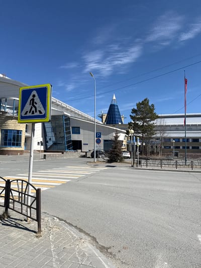 The image depicts a street scene in Beloyarsky, Russia. A crosswalk sign is prominent in the foreground, with a pedestrian crossing extending across the road. A building with a pointed, glass-covered roof is behind the sign. A street light is positioned nearby, reaching upwards to the sky. The sky is clear blue with wisps of clouds. A parking sign is visible near the building. Further in the background, a multi-story building is visible behind a tree. A red flag is visible near the top of the multi-story building. The street is paved with asphalt and a metal barrier is present on the side of the road.