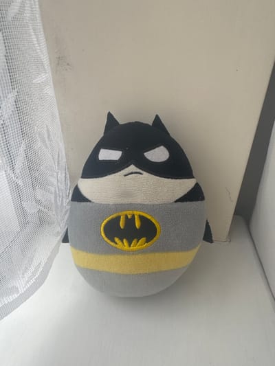 A Batman plush toy leans against a white wall and ceiling in a room. Part of a white, mesh-like object is in the foreground, obscuring the lower portion of the image. The toy has a black and white face and gray body with a yellow stripe featuring the Batman logo. The location in Welshpool, United Kingdom is undetermined.
