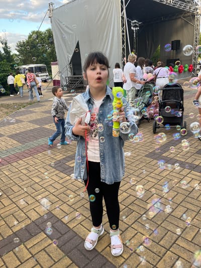 In the city of Malaryta, Belarus, a young girl in a denim jacket and black leggings stands on a brick-paved area, holding a bubble gun. She is surrounded by numerous soap bubbles floating in the air. Behind her, a white stage with a dark backdrop is set up, and people are gathered, some pushing strollers. Other individuals are visible in the background, including children and adults, and a white bus is parked to the left of the scene. The environment appears to be an outdoor public space, possibly a park or event area, with trees and greenery surrounding the stage. The sky above is partly cloudy.