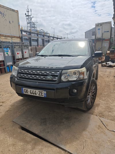 A black Land Rover Discovery is parked outdoors on a dirt surface in an industrial or port area. The vehicle is positioned facing the camera, with its front bumper slightly elevated by a metal plate on the ground. To the left of the car are several tall metal racks filled with blue and red gas cylinders, some of which are labeled with "AIR LIQUIDE" and hazard symbols. Behind the car, a large ship or barge with a complex superstructure is docked. In the background, there are industrial buildings and shipping containers, one of which is white with the number "5" printed on it. The sky is cloudy, suggesting overcast weather. The license plate on the car reads "GK-444-ZE 62", indicating it is registered in France. The overall scene depicts an industrial waterfront environment under a cloudy sky.