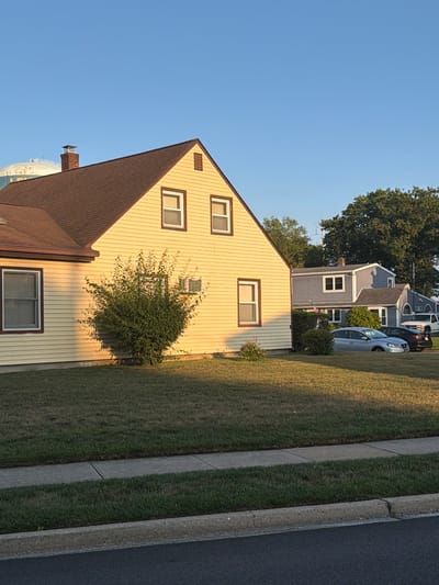 The image shows a residential street in Levittown, United States of America, on a clear day. The foreground features a lawn with neatly mown grass bordered by a sidewalk and a street. On the left, a pale yellow house with a brown roof dominates the frame, with its side facing the viewer. A large evergreen shrub is planted in front of the house. In the background, a grey house with a white trim and a dark roof is visible, with a white pickup truck parked in front of it. The sky is a clear blue, with a large water tower in the far distance. The lighting suggests it is either early morning or late afternoon, as the sun casts long shadows across the lawn and the side of the yellow house. No people are visible in the image.