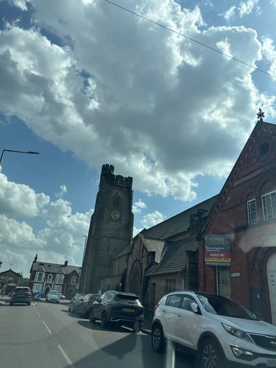Captured from a vehicle's vantage point, the image depicts a street scene dominated by a historic church tower. The tower, constructed from gray stone, features a clock face and ornate architectural details. Adjacent to the church is a red brick building with signage indicating a "Redevelopment Opportunity," including a phone number. Cars are parked along the street, including white and black vehicles. The sky is filled with a mix of blue sky and large cumulus clouds. The presence of the church tower and building styles suggest the location is in Nottingham, United Kingdom.