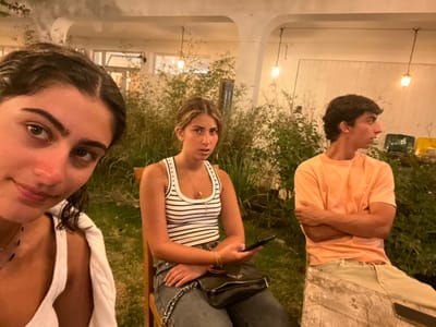 The image captures three individuals in an illuminated outdoor setting during the evening or night in Aljezur, Portugal. The scene appears to be a patio or garden area adjacent to a light-colored building.

On the far left, partially visible in the foreground, is a young woman with dark, curly hair, wearing a white sleeveless top with a white garment draped over her left shoulder and a dark beaded necklace. She faces the camera directly with a slight smile.

In the center, a young woman with light brown hair is seated on a wooden chair. She wears a white and navy blue striped sleeveless top, dark trousers, and a thin gold chain necklace. A black handbag with a silver chain strap rests on her lap, and she holds a dark-colored smartphone in her right hand. Her gaze is directed towards her left.

On the right, a young man with dark, curly hair is seated on a large, cut log or wooden bench. He wears a plain orange short-sleeved t-shirt and light-colored trousers. His arms are crossed over his chest, and he is looking towards his right.

The background features dense green foliage or tall grasses, with a light-colored building structure visible behind it, including what appears to be a window or doorway and an arched overhead section. Several warm-toned pendant lights or string lights are suspended from an unseen structure, providing the primary illumination. The ground beneath the subjects appears to be natural earth or sparse grass. Faintly discernible colored objects are present in the darker background behind the man on the right.