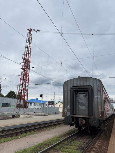 This is a photograph taken at a train station in Povorino, Russia, under a cloudy sky. The foreground shows railway tracks and a portion of a train car, viewed from the rear. The train car is dark gray with red trim and visible details such as lights, coupling mechanisms, and Cyrillic lettering indicating its origin and destination. To the left of the train, a red metal pylon stands tall, supporting overhead electrical wires for the train. Behind the pylon, a building with a bright blue roof and white walls is visible. Further to the left, a gray metal fence encloses an area with some industrial-looking structures. The overall scene depicts a typical railway environment on an overcast day.