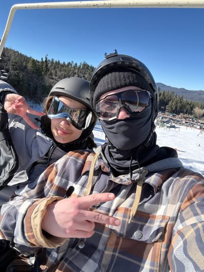 A selfie captures two people wearing ski helmets and goggles, flashing peace signs. The background reveals a blue sky, trees, and snow-covered ground. The individual in the foreground has silver reflective goggles, while the other is wearing a black face covering. The person closest to the camera is wearing a grey jacket. The other person is wearing a plaid jacket. The landscape features mountains and buildings. They appear to be on a ski lift.