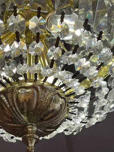 The image shows a close-up view of a chandelier with many faceted crystals and brass accents. The crystals are arranged in a dense cluster, catching the light and creating prismatic effects. A decorative brass element with intricate carvings is visible at the bottom of the frame. The background is dark and out of focus, emphasizing the illuminated details of the chandelier. There are no people, actions, or interactions depicted. The image appears to be a still-life detail shot and does not provide any information about its location in Donegal, Ireland, or any other city.