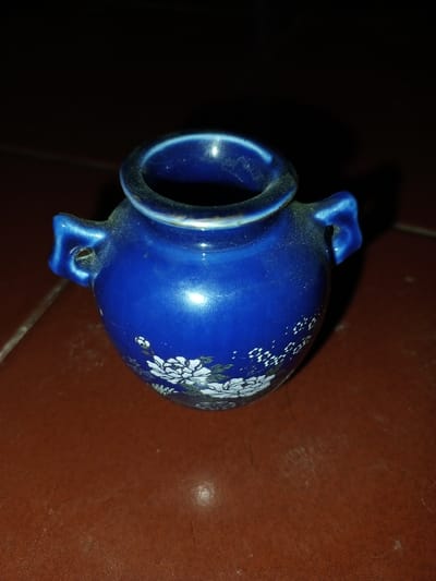A small, dust-covered, cobalt blue ceramic vase is centrally positioned on a reddish-brown, textured surface. The vase features a flared rim, a constricted neck, a bulbous body, and two small loop handles on opposing sides of the upper body. White floral patterns, consisting of blossoms and leaves, decorate the lower half of the vase's body. Smaller, indistinct white patterns are visible above the main floral design. The background is predominantly dark, with a shadow cast to the right of the vase. No individuals, actions, or interactions are depicted. The image provides no visual information to ascertain its location within the city of Longmiang, Laos.
