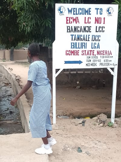 A young person walks past a white sign that reads "Welcome to ECWA LC No 1, Banganje LCC, Tangale DCC, Billiri LGA, Gombe State, Nigeria." The sign also lists service times: "Sun School 7:30-8:30 am, Sun Service 8:30 am, Mid-Week Prayer 4-5pm." The location is confirmed as Billiri, Nigeria, based on the sign's inscription. The environment appears to be a rural or semi-rural area, given the unpaved ground and the presence of a ditch or drainage area.