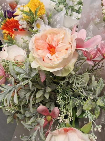 This media file is a close-up, eye-level, medium shot of a bouquet of artificial flowers. The flowers are arranged in a lush and vibrant display, featuring a large, multi-layered peach-colored rose as the focal point. Surrounding the rose are other blossoms in shades of pink, yellow, orange, and white, with small, delicate white filler flowers and green foliage. The bouquet is partially wrapped in clear cellophane that is decorated with a pattern of small white flowers and hearts. The background is out of focus and appears to be a solid dark surface. There is no visible text in the image. The lighting is bright and even, suggesting an indoor setting, possibly a shop or a display. The overall impression is one of a decorative and aesthetically pleasing floral arrangement, likely intended for gifting or home decor. No people are visible, and no specific activity or event is taking place, other than the presentation of the bouquet. The scene is static and focused solely on the artificial flowers.