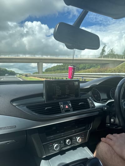 This media file captures the interior of a car driving on a highway. The perspective is from the driver's seat, looking forward. Visible subjects include the dashboard with its infotainment system displaying radio stations and settings, a rearview mirror, and the steering wheel. A person's arm and hand are visible, suggesting the driver.
The scene is set on a highway, indicated by the road, guardrails, and overpasses visible through the windshield. The weather appears to be partly cloudy, with a mix of blue sky and white clouds. The lighting suggests it is daytime, likely late morning or early afternoon.
A notable detail is a Coca-Cola branded air freshener hanging from the rearview mirror. The infotainment screen displays several radio station presets, including RTE R1, RTE 2FM, RTElyric, RTE RnaG, Today FM, and Newstalk, all common in Ireland. The time displayed on the screen is 3:18. The dashboard also shows climate control settings and other vehicle functions. The overall impression is a mundane journey on a highway, with the driver presumably engaged in listening to the radio.