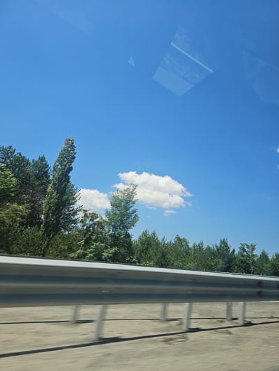 The image captures a scene from a moving vehicle, likely on a highway, with a bright blue sky and scattered white clouds. In the foreground, a silver guardrail runs horizontally across the frame, its metallic surface reflecting the ambient light. Beyond the guardrail, a line of lush green trees forms a dense treeline, with various types of coniferous and deciduous trees visible. The ground beneath the guardrail appears to be a sandy or gravelly surface with a slight blur, indicating motion. In the sky, a few fluffy white clouds are present, and there are subtle reflections on the vehicle's window, suggesting the photograph was taken from inside. The general setting appears to be a rural or semi-rural area, possibly bordering a highway, near Staro Selo, Bulgaria, judging by the presence of typical vegetation and road infrastructure found in such locations. There are no people or discernible actions taking place.