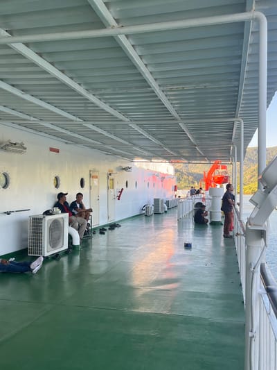 The image depicts the deck of a ferryboat, likely in the vicinity of Batas Desa Skotong Timur, Indonesia, given the lush green hills visible in the background. The deck is a light green color and is partially covered by a white metal canopy. Several people are present on the deck, some sitting and conversing, others standing and looking out at the water.

In the foreground, a person lies on their side, resting. To the left, near a white air conditioning unit, two men are seated, one wearing a black baseball cap and a red jacket, the other in a dark t-shirt and shorts. Further down the deck, several pairs of sandals are neatly placed on the floor.

Mid-deck, a red lifeboat davit system is visible. A person in a dark shirt and patterned pants stands near the railing, looking out towards the water and the distant hills. Other individuals are seen scattered on the deck, some appearing to be waiting or relaxing. The water is calm and reflects the light from the sky. The overall scene suggests a relaxed atmosphere, typical of ferry travel.