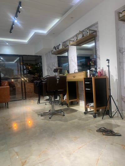 The image displays the interior of an unoccupied hair salon or barber shop located in Lauro de Freitas, Brazil, appearing to be captured during evening or nighttime hours due to the dark exterior visible through the windows. The establishment is well-illuminated by a black track lighting system with multiple spotlights mounted on the white ceiling, complemented by recessed lighting in the ceiling alcoves. The floor is covered in polished light-toned marble-patterned tiles, which reflect the interior lights.
The walls are predominantly white, with sections featuring a grey and white marble-like pattern serving as a backdrop for the workstations. Two dark brown, adjustable hydraulic salon chairs with chrome bases are visible, one positioned centrally in the foreground-midground with a dark fabric draped over its back. To the far left, a brown upholstered armchair is partially visible near the main entrance.
Two salon workstations are present, each equipped with a large rectangular mirror and a light wooden console featuring gold-colored metal accents. The console in front of the central chair includes a single drawer. Above each mirror, a gold-framed shelf holds various hair product bottles and containers; the right shelf also displays decorative barber tools. Two mobile salon carts, constructed with light wooden frames and black panels, are on casters. One cart, adjacent to the central workstation, has multiple drawers/shelves and holds numerous product bottles on its top surface.
A black tripod stand is positioned upright on the right side of the image. A pair of black flip-flops with patterned straps lies on the floor in the lower right corner. The left side of the room features large glass doors and windows with black frames, providing a view of the dark street outside, where vehicle lights are discernible. Reflections from both interior and exterior light sources are visible on the polished floor and within the mirrors.