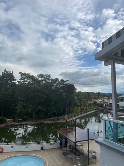 This image captures an outdoor scene from an elevated perspective, likely in a resort or residential complex in Floridablanca, Colombia. The foreground on the lower left shows a light blue swimming pool with a white curved edge and a red lifebuoy attached to a white railing. Adjacent to the pool area, to the right, is an outdoor dining or lounge space with a large, dark, rectangular canopy supported by metal poles. Beneath this canopy, several dark metal tables and chairs are arranged. Two individuals are standing near the water's edge below the canopy; one wears a blue shirt, and the other wears a dark shirt.
A long, narrow body of green-tinted water, appearing to be a canal or pond, extends across the midground. Its surface reflects the sky and surrounding foliage. A wooden walkway or bridge with wooden railings spans portions of this water body and runs along its far bank. Several small figures, appearing to be pedestrians, are visible walking on this walkway in the distance.
The background on the left is dominated by a dense forest of tall, green trees. To the right of the water body, beyond the walkway, several residential buildings with distinct white and reddish-brown rooftops are visible, indicative of a developed area. Distant mountains or hills are faintly discernible on the horizon beneath a sky characterized by a mixture of white and grey clouds with patches of blue. On the far right, part of a building structure with a white facade and a top section featuring horizontal green and white stripes is visible.