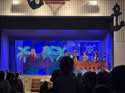 A stage is set for a children's play, likely a pirate-themed performance, in Ufhusen, Switzerland. The backdrop depicts a vibrant, colorful scene with palm trees, a volcano, and choppy blue waves, suggesting a tropical island setting. Prominently featured is a large, cardboard ship labeled "ESMERALDA" with a cannon. Several children, dressed as pirates and wearing hats and vests, are on the ship's deck, some holding microphones. Behind them, a Jolly Roger flag is displayed.
In the foreground, the audience is visible, with the backs of their heads and shoulders filling the lower part of the frame. Their attire suggests it is likely daytime, possibly an evening performance given the stage lighting. The atmosphere appears to be one of anticipation and enjoyment, with the focus on the young performers. Above the stage, decorative black anchors hang from the ceiling, reinforcing the pirate theme. The overall impression is a community event, likely a school play, filled with youthful energy and a touch of theatrical magic.