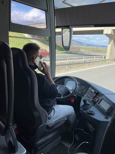 The image depicts the interior of a moving bus or coach, viewed from a passenger's perspective looking forward and to the right, showing the driver and the external environment.

**Scene Overview:**
The scene is set inside the driver's cabin of a large passenger vehicle, likely a Yutong bus, traveling on a multi-lane highway. The weather is clear with scattered clouds. The location is a highway or major road within or near the Градска општина Црвени Крст, Serbia, characterized by concrete overpasses and distant hills.

**People:**
One adult male, presumed to be the driver, is visible from the back and right side. He has short, dark hair and is wearing dark sunglasses pushed up on his head or just above his eyes. He wears a dark long-sleeved shirt or jacket and light grey trousers, possibly jeans. He is seated in a dark grey driver's seat with a seatbelt fastened. His left hand rests on the steering wheel, while his right hand holds a light-colored smartphone (appearing to be an iPhone with a multi-lens camera system) to his right ear, indicating he is engaged in a phone call while operating the vehicle.

**Vehicle Interior:**
The driver's compartment features a black steering wheel with a silver Yutong logo. A red heart-shaped ornament hangs from the steering column. The dashboard is dark grey, equipped with an instrument cluster displaying gauges, a radio/media unit, control buttons, and a digital screen. A manual gear shifter with a black knob is positioned to the driver's right. White charging cables are visible on the floor and dashboard area, with one connected to a USB adapter. German-language warning labels are present: "Bitte während der Fahrt nicht mit dem Fahrer sprechen." (Please do not talk to the driver while driving.) on a side window frame, and "Rollo elektrisch angetrieben! Nicht ziehen!!!" (Electric blind! Do not pull!!!) above the external side mirror.

**Exterior Environment:**
Through the bus windows, a highway is visible with asphalt pavement, white lane markings, and metal guardrails on the shoulder. The bus is passing under a massive, light grey concrete bridge or overpass with visible support pillars. Beyond the highway, green grassy embankments with sparse trees ascend, leading to rolling hills and mountains in the distance under a partly cloudy blue sky. A small, older-model red car (resembling a Zastava 750 or Fiat 600) is parked or stationary on the grassy area next to the highway, under the concrete overpass, on the left side of the bus's direction of travel.