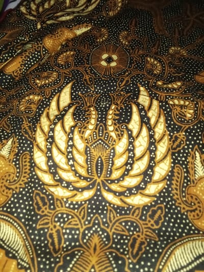 The image presents a close-up view of a dark-colored textile fabric patterned with traditional Indonesian batik designs. The dominant color scheme includes a black or very dark brown background, complemented by golden-brown and off-white motifs. A prominent, centrally located design depicts a highly stylized mythological bird figure, suggestive of a Garuda or similar creature, rendered with a crown-like head, intricate feather details, and symmetrical wings composed of layered, pointed forms. Surrounding this central motif are various smaller floral elements, swirling vine patterns, and dotted textures, all executed in contrasting golden-brown and off-white. The fabric exhibits subtle folds and creases inherent to textile materials. This fabric is located in Umsobomvu Local Municipality, South Africa.