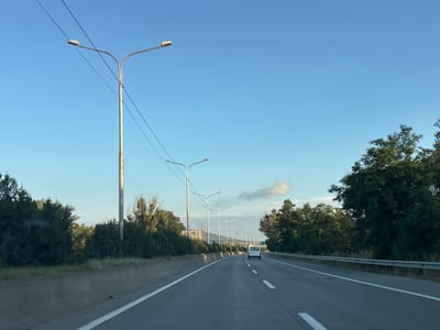 The image depicts a straight, multi-lane highway in Changilari, Georgia, under a clear blue sky with a few wispy clouds. The road is paved and marked with white lines. On the left side of the road, there is a concrete barrier, followed by vegetation and tall streetlights with multiple lamps. The streetlights are also present on the right side of the road, spaced at intervals along the highway. Dense green trees line both sides of the road, creating a naturalistic border. In the distance, a white van is traveling on the highway. The overall scene suggests a rural or suburban highway environment.