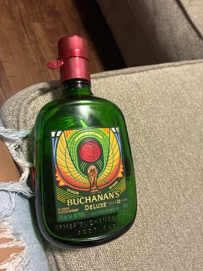 A green glass bottle of Buchanan's Deluxe Scotch Whisky, featuring a FIFA World Cup 2026™ Edition label, is held by an unseen individual. The label is circular with a red seal in the center, surrounded by a colorful design of wings and a gold trophy. Text on the label includes "UN BALÓN," "BUCHANAN'S," "DELUXE AGED 12 YEARS," "BLENDED SCOTCH WHISKY," "40% alc./vol. (80 PROOF)," and "750 ml." The bottle has a red, textured cap with a tamper-evident seal. The bottle is resting on a textured grey surface, possibly a sofa or upholstered chair. The lower portion of the image shows ripped denim fabric, likely from jeans worn by the person holding the bottle. The background is blurred and appears to be indoors, with hints of wood flooring and darker shadows suggesting a dimly lit environment. There is no indication of the city of Dallas, United States of America in the image.