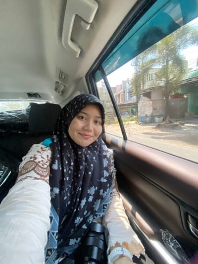 A young Indonesian woman wearing a black and white floral hijab and a cream-colored blouse with brown detailing is taking a selfie from the back seat of a car. The car's interior is visible, including the ceiling with a grab handle and air vents, and the brown leather door panel. Outside the car window, buildings and trees are visible, suggesting an urban or suburban setting. The woman is smiling at the camera. The location is identified as Mandar Sari, Indonesia.